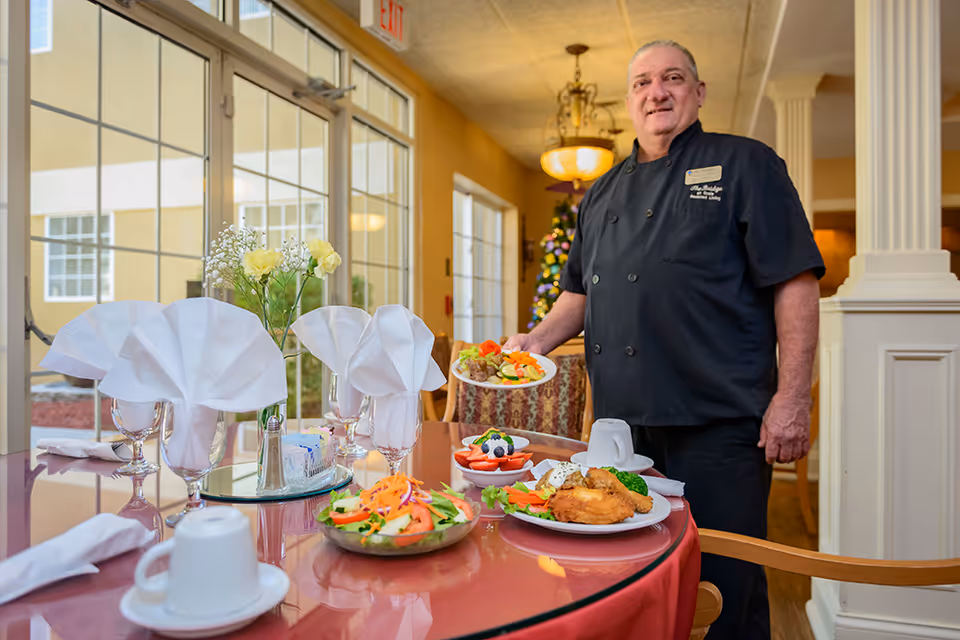 A chef stands in a dining room beside a table set with plated meals, folded napkin-stemmed glasses, and a vase of flowers.