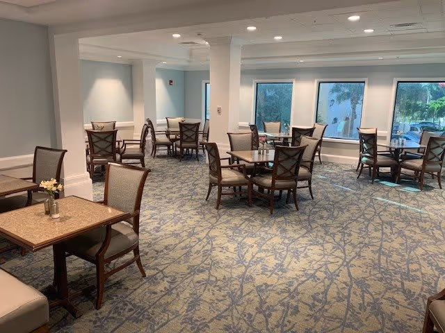 A spacious dining room with multiple square tables and cushioned chairs arranged neatly on a patterned carpet. Large windows allow natural light to brighten the room, and the walls are painted in a soft neutral color.