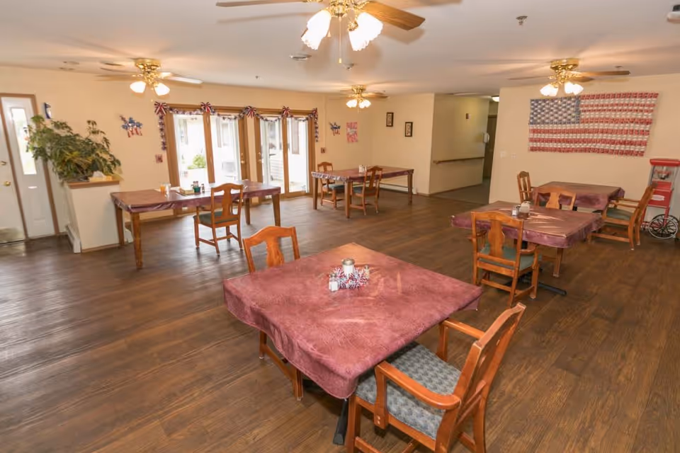 A dining room in Scandinavian Court Assisted Living with several wooden tables covered with maroon tablecloths and wooden chairs. The room has wooden flooring, ceiling fans with lights, and patriotic decorations including an American flag made of small items on the wall. There are large windows and a door letting in natural light.