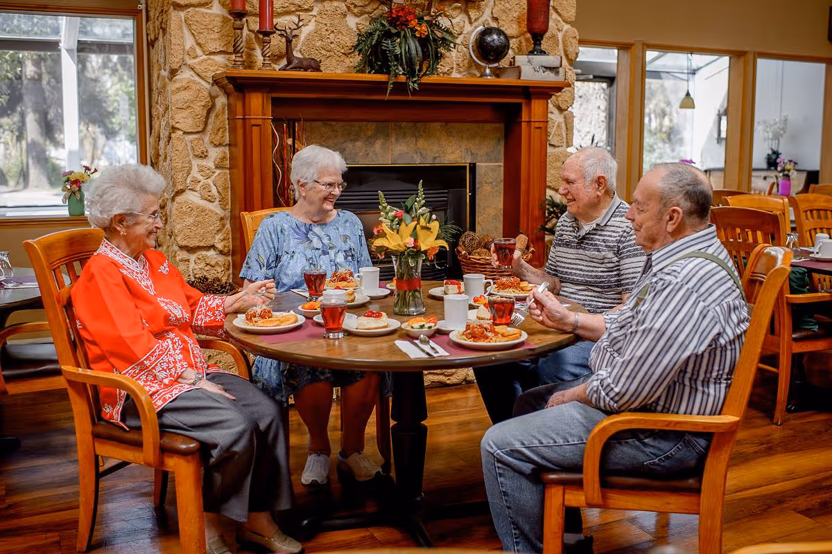 Four elderly people sitting around a round wooden dining table in a cozy room with a stone fireplace, enjoying a meal together with plates of food and glasses of red drink, smiling and engaging in conversation.
