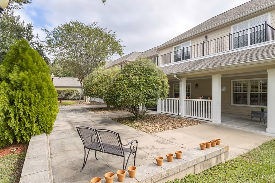Outdoor patio area at Brookdale Spring Shadows featuring a paved walkway, a black metal bench, several small terracotta pots lined up on a low wall, green bushes, trees, and a two-story building with a covered porch and balcony.