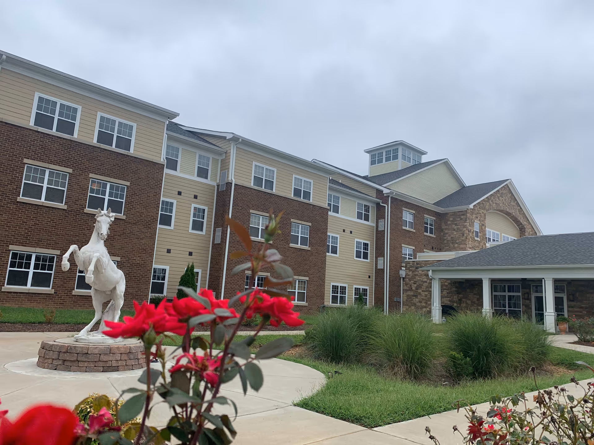 Exterior view of a senior living facility building with multiple windows, a covered entrance, and a white horse statue on a circular brick pedestal in the landscaped front area with red flowers and green shrubs.