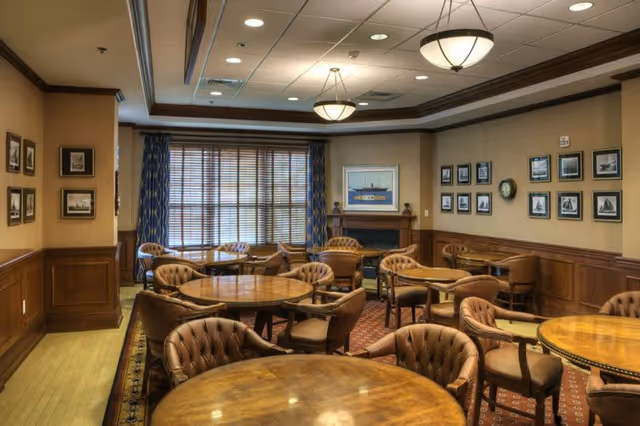 A cozy senior living common area with several round wooden tables surrounded by cushioned chairs. The room features wood paneling on the walls, framed pictures, a fireplace, and large windows with blue patterned curtains allowing natural light to enter.