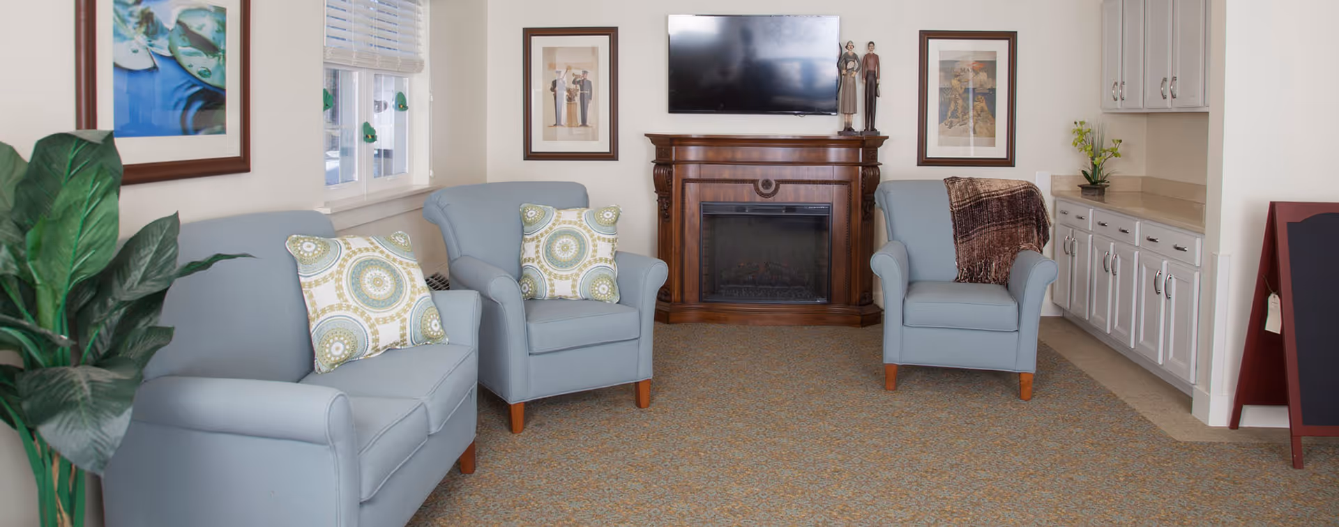A cozy living room with three light blue armchairs, two with patterned cushions and one with a brown throw blanket. A wooden fireplace with a flat-screen TV mounted above it is centered on the wall. There are framed pictures on either side of the fireplace, white cabinetry with a countertop on the right, and a large green plant on the left near a window with white blinds.
