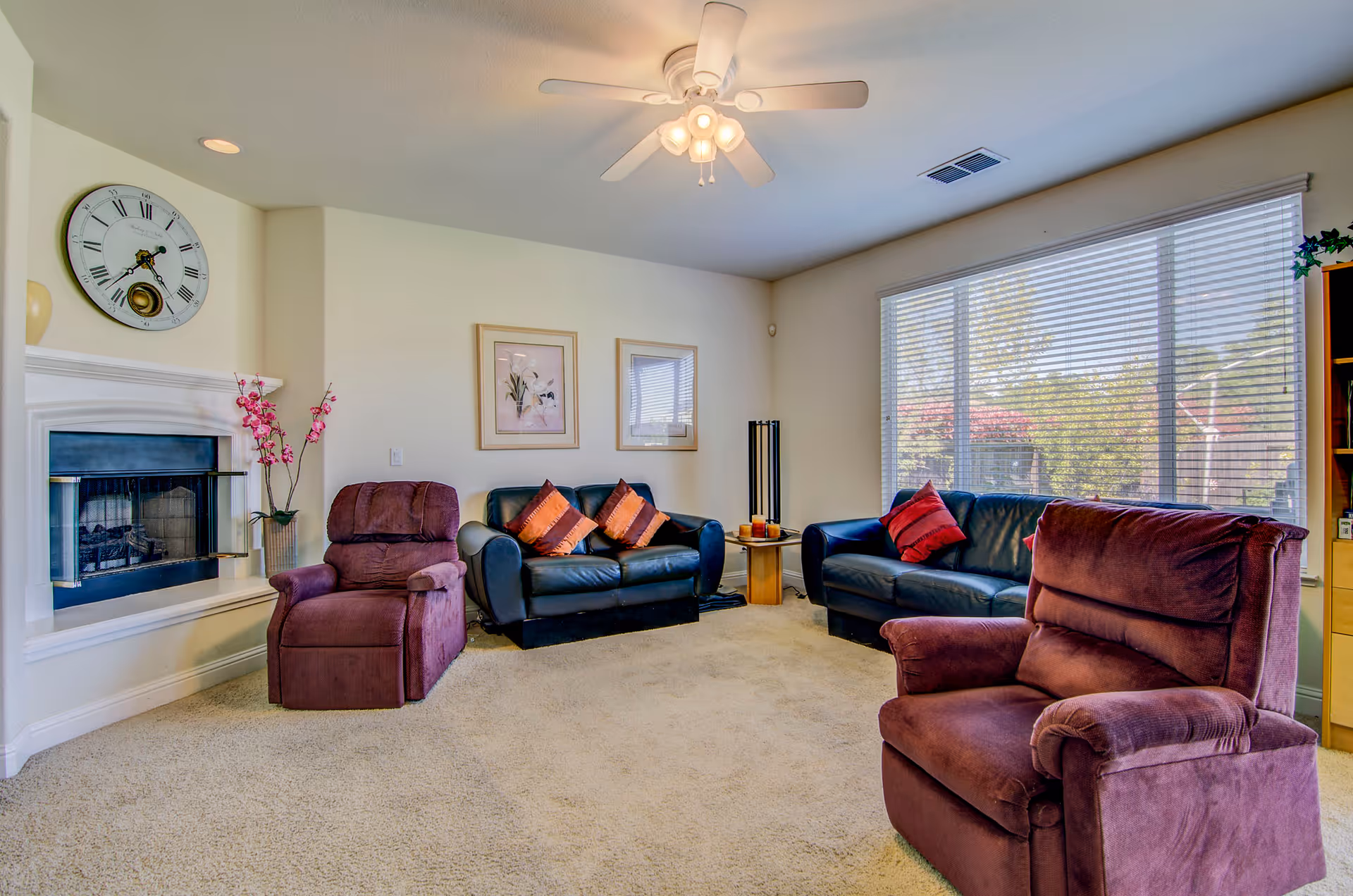 A cozy living room with beige carpet, two purple recliners, two black leather sofas with red and orange cushions, a white ceiling fan with lights, a large window with blinds letting in natural light, a fireplace with a large clock above it, and framed floral artwork on the wall.