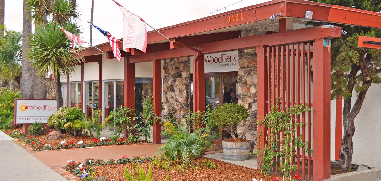 Exterior view of a single-story building with stone and red wooden accents, surrounded by landscaped plants and flowers. The building has a sign that reads 'WoodPark Specialty Elder Care' and the address number 3121 is visible on the roof edge. Flags are displayed above the entrance.