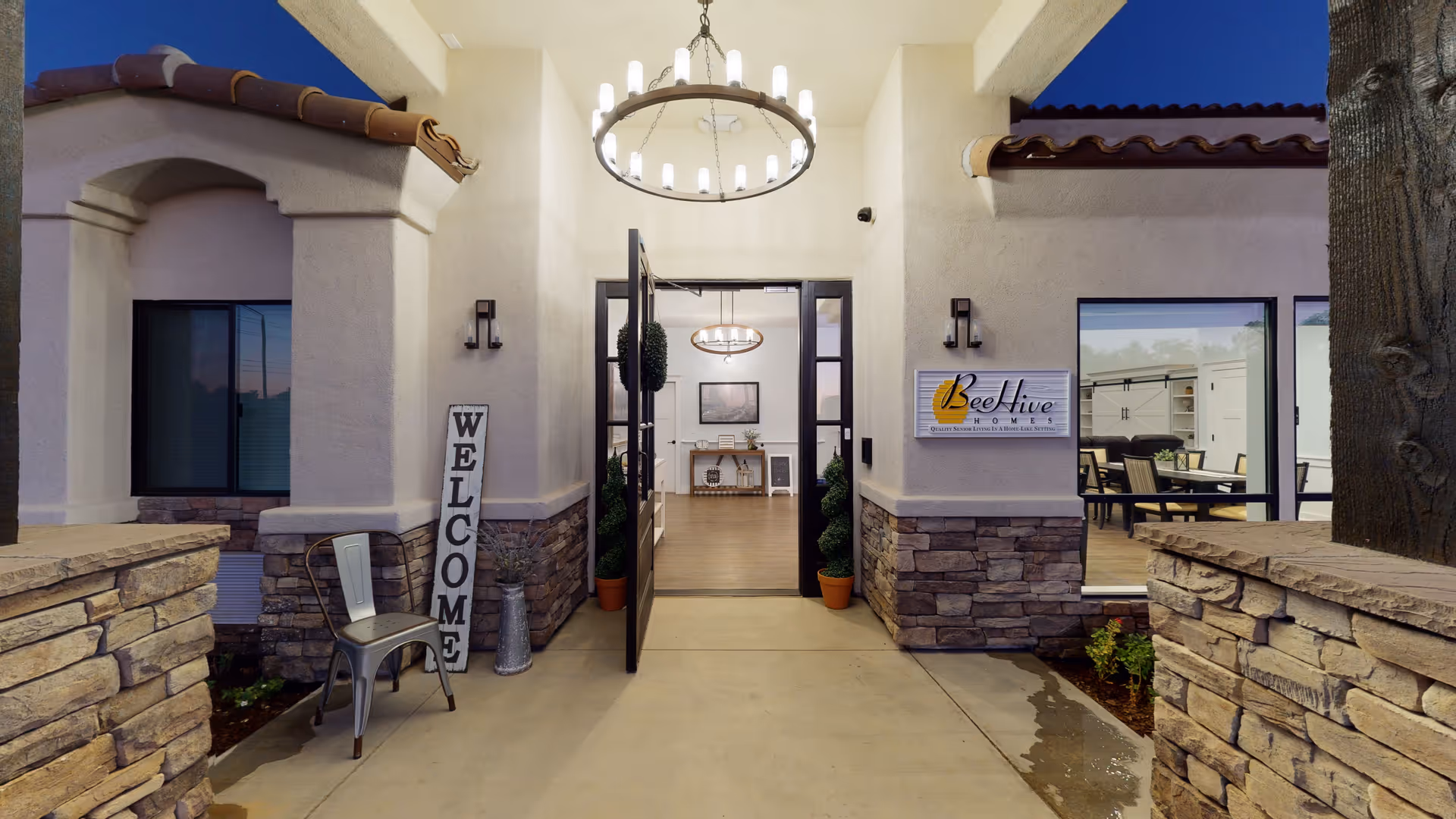 Entrance to BeeHive Homes Assisted Living facility at dusk, featuring an open glass door, a 'WELCOME' sign leaning against the wall, a metal chair, stone and stucco exterior walls, and a chandelier hanging from the ceiling. Through the door, an interior space with wooden flooring and furniture is visible.