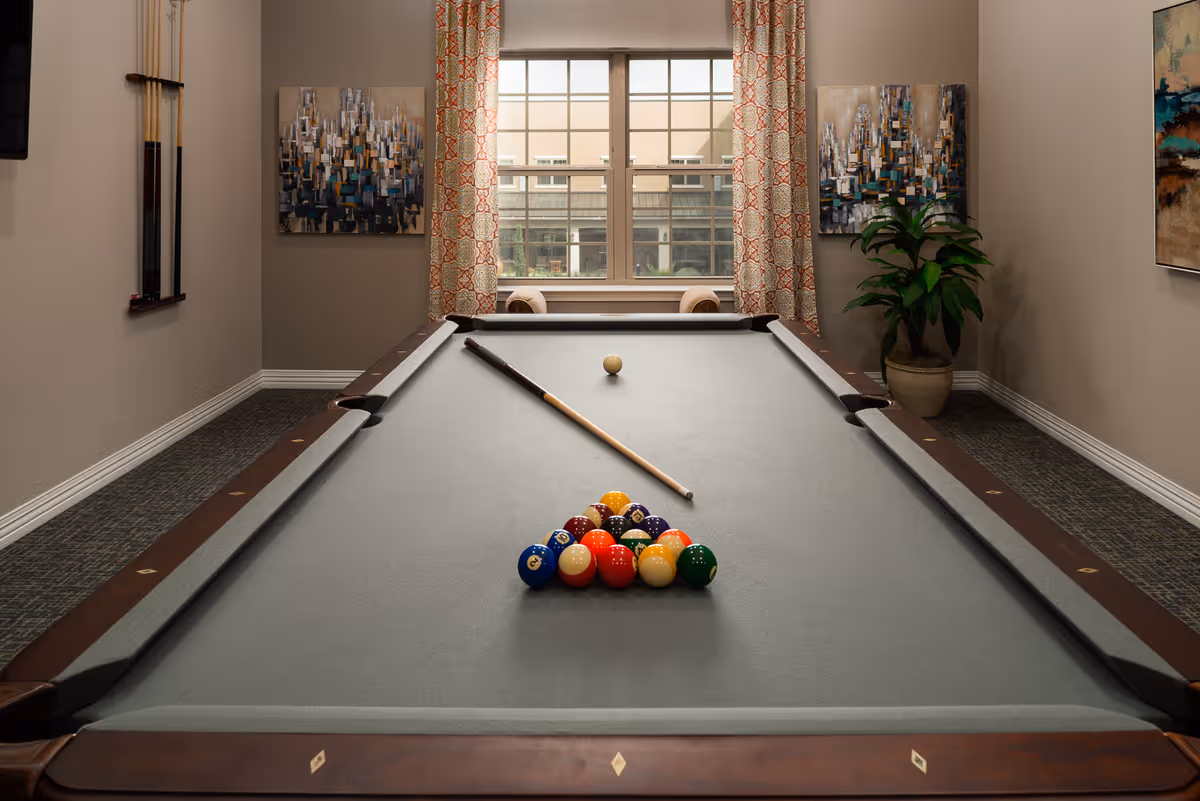 A pool table with a full rack of colorful billiard balls and a cue stick resting on the table in a well-lit room. The room has a window with patterned curtains, two abstract paintings on the walls, a potted plant, and a rack holding additional pool cues.