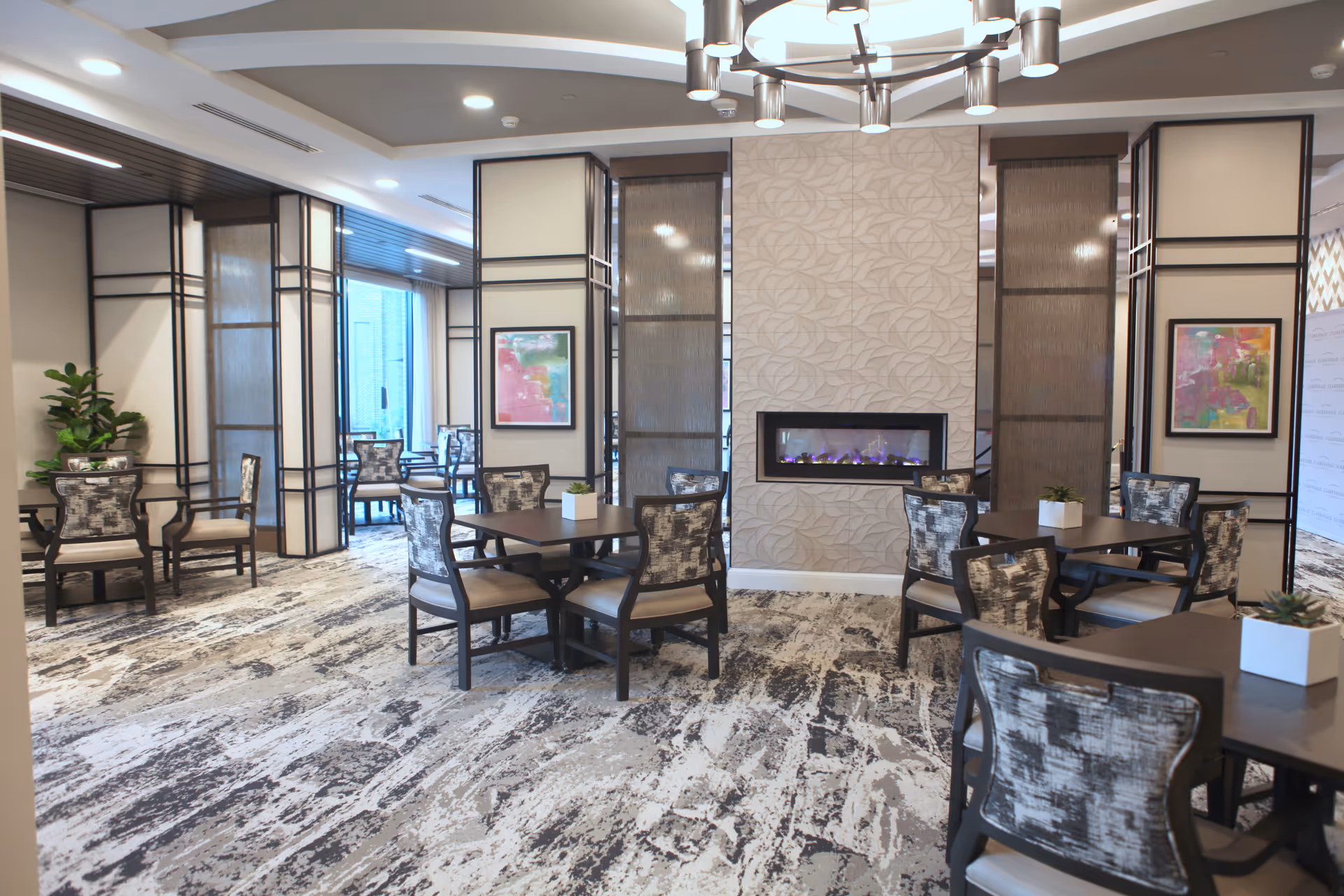 A modern dining area in a senior living facility with multiple square tables surrounded by patterned cushioned chairs. The room features a textured wall with a built-in electric fireplace, abstract artwork on the walls, and large windows letting in natural light. The floor has a patterned carpet and there are decorative plants on some tables.