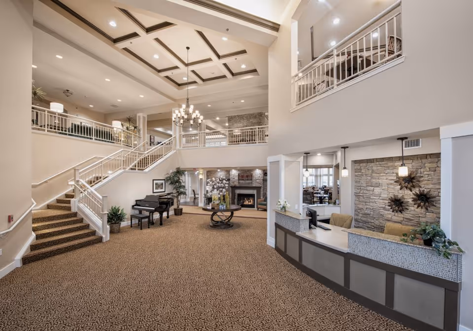 Spacious and elegant interior of a senior living facility featuring a large open lobby with a patterned carpet, a grand piano, a round table with floral arrangements, a stone fireplace, and a reception desk with two chairs. The area has high ceilings with recessed lighting and a chandelier, a staircase leading to an upper level with railings, and decorative wall art.