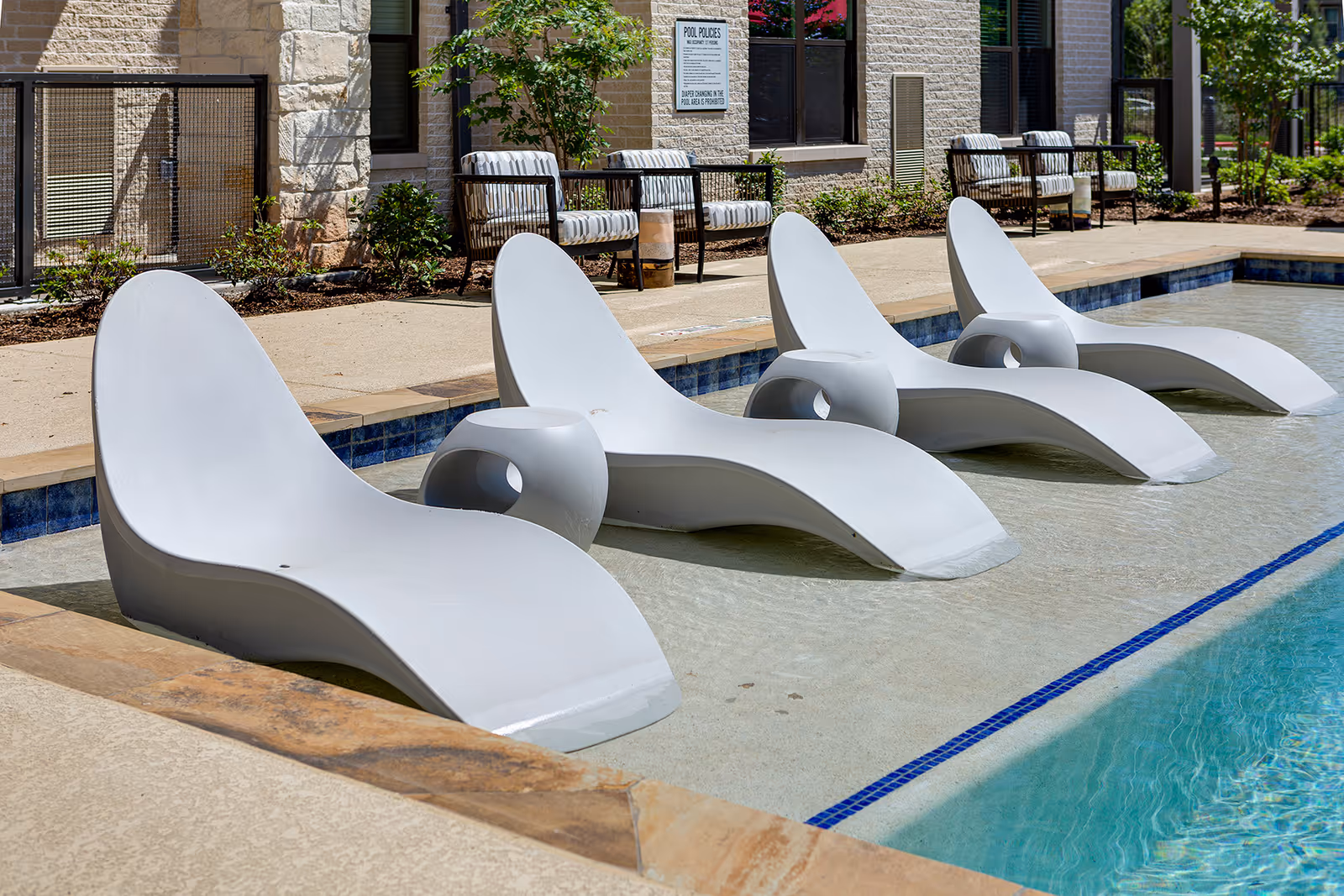 Four modern white lounge chairs partially submerged in the shallow end of a swimming pool with a small round table between each pair. In the background, there are cushioned chairs and a stone building wall with windows and a pool rules sign.