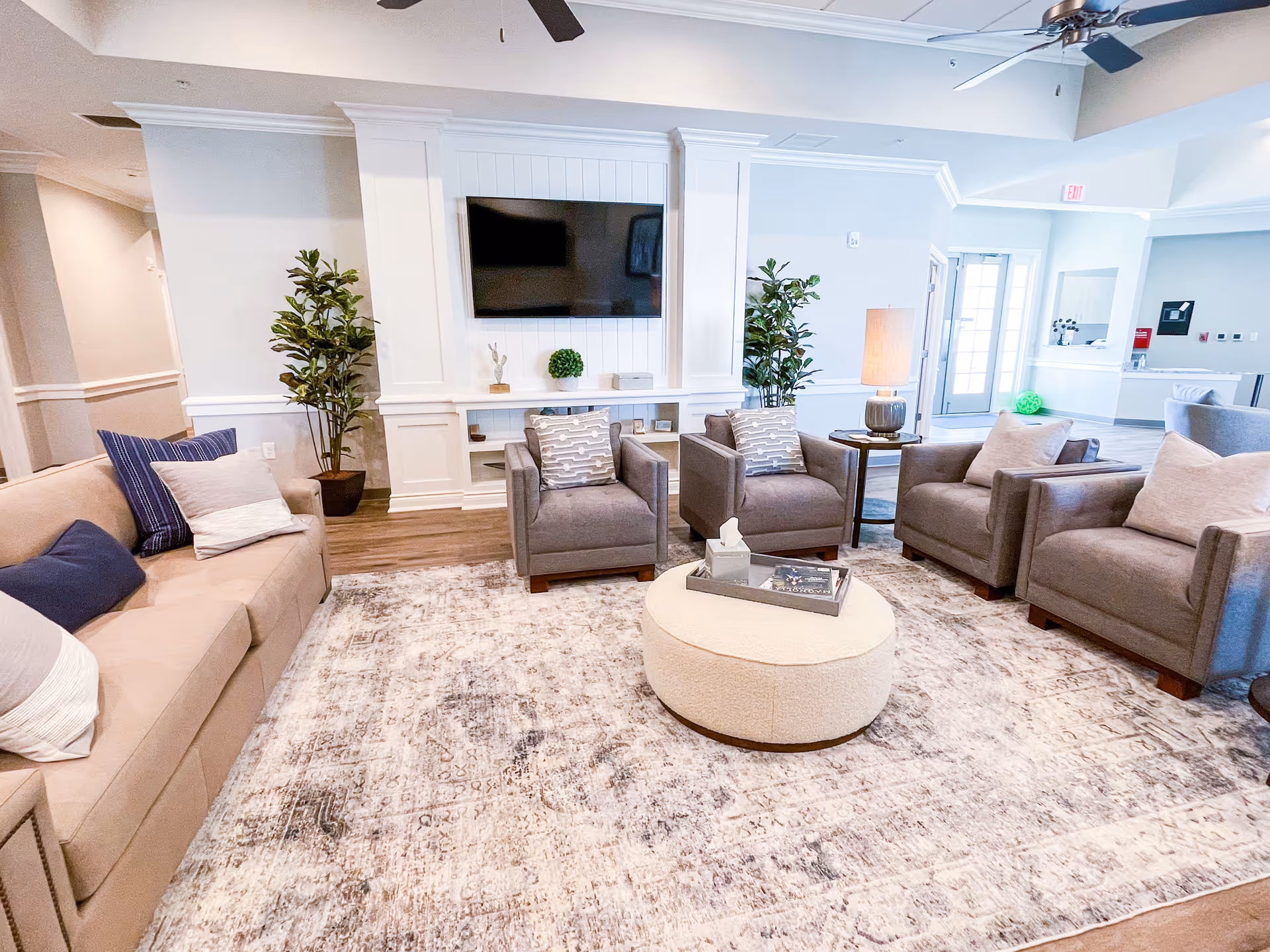 A bright and spacious senior living facility common area with a beige sofa and four gray armchairs arranged around a round ottoman on a patterned rug. A flat-screen TV is mounted on a white paneled wall, flanked by two tall green plants. There is a side table with a lamp and a hallway leading to other rooms in the background.