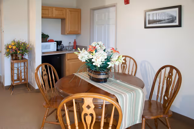 Dining area with a round wooden table and chairs, a floral centerpiece, and a small kitchenette in the background.