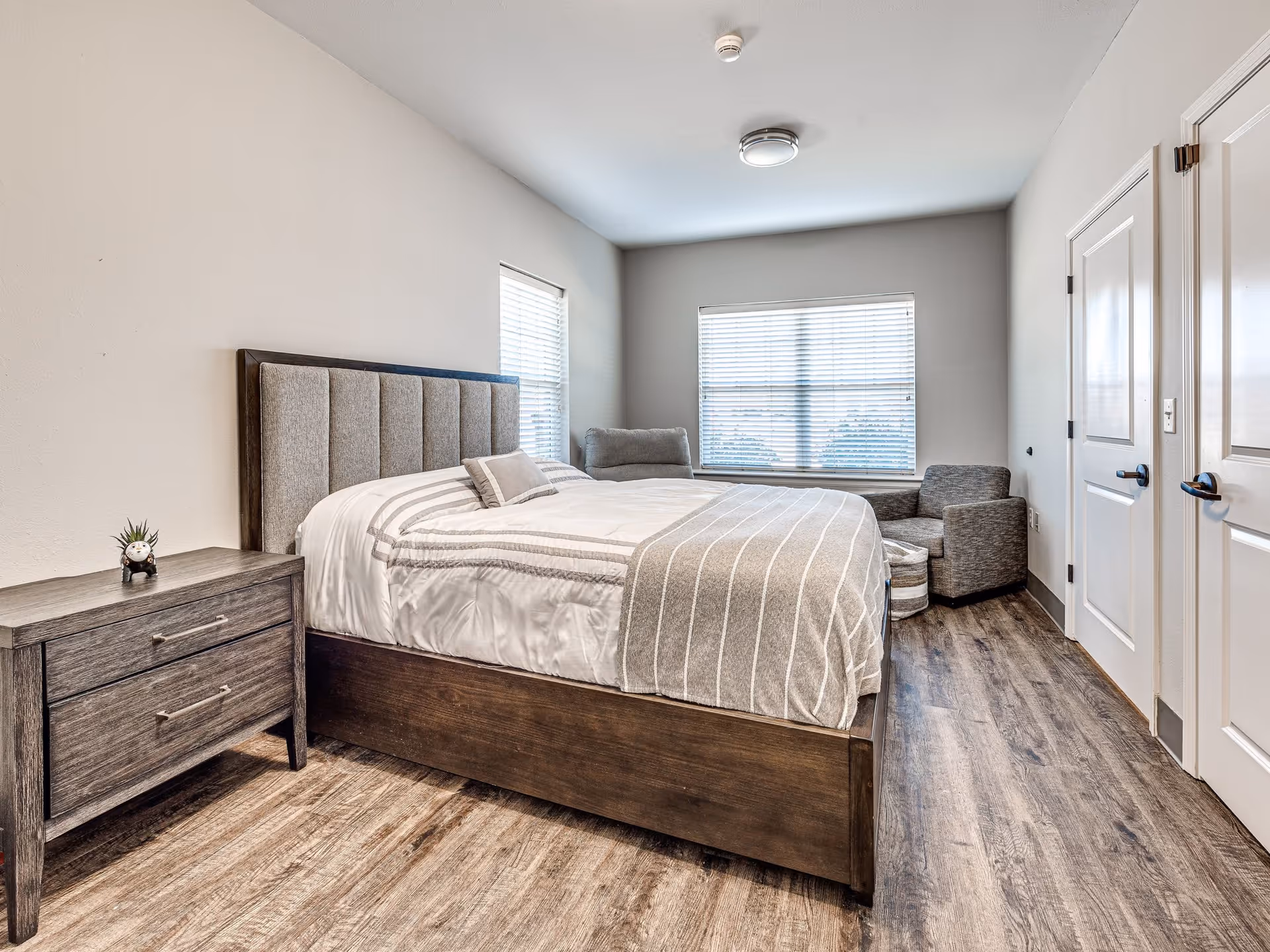 A bright bedroom with a large bed featuring a padded headboard, white and gray bedding, a wooden nightstand with a small plant and clock, two gray armchairs near a window with blinds, and two closed white doors on the right side.