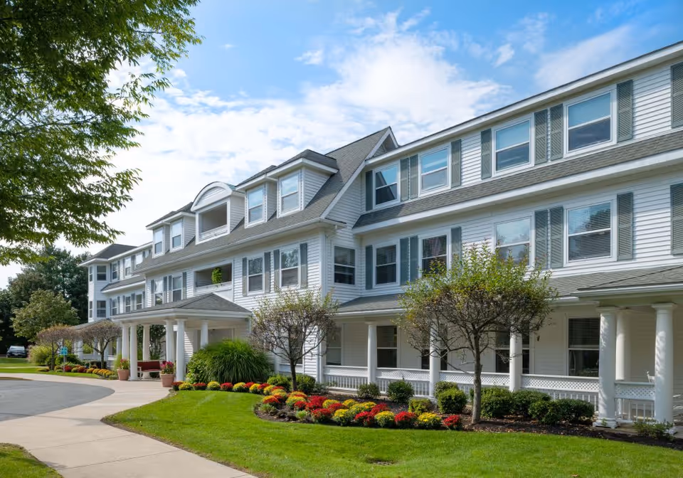 Exterior view of a large, white senior living facility building with multiple windows and a covered entrance. The building is surrounded by well-maintained landscaping including green grass, small trees, and colorful flower beds under a partly cloudy sky.
