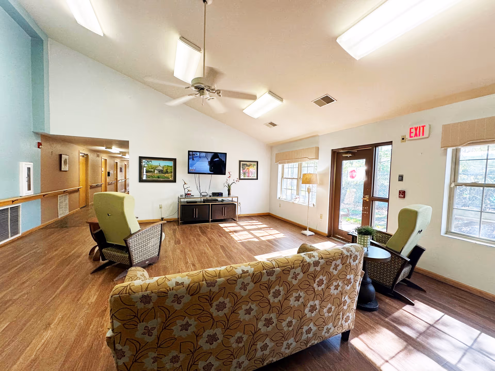 A bright and spacious living room area in a senior living facility with a floral patterned sofa, two green armchairs, a small round table with a plant, a TV mounted on the wall, framed pictures, large windows letting in natural light, and a door with an exit sign above it.