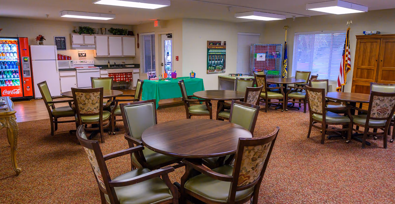 A communal dining area with several round wooden tables and chairs with green cushions and floral backs. In the background, there is a kitchen area with white cabinets, a refrigerator, a stove, and a Coca-Cola vending machine. A green tablecloth-covered table with craft supplies is also visible. The room has carpeted flooring, windows with blinds, and flags in the corner.