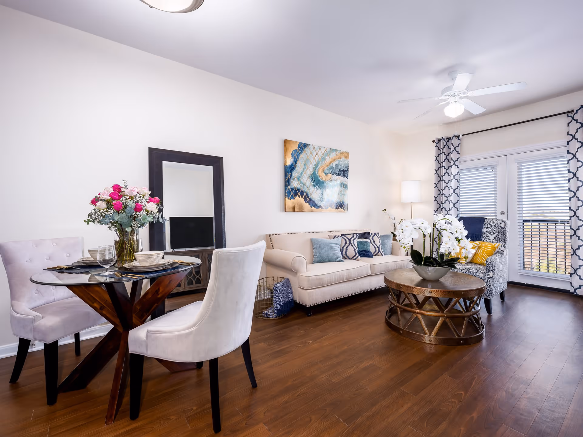 A bright living room with a beige sofa adorned with blue and patterned cushions, a round wooden coffee table with a white orchid centerpiece, a patterned armchair with a yellow pillow, and a floor lamp. There is a small round dining table with two upholstered chairs, set with plates and glasses, and a vase of pink and white flowers. The room has wooden flooring, a large mirror leaning against the wall, abstract artwork above the sofa, and French doors with patterned curtains letting in natural light.