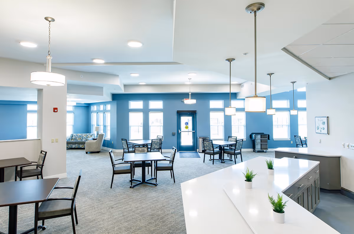 A bright and spacious common area in an assisted living facility with multiple tables and chairs arranged for dining or socializing. The room features blue accent walls, large windows letting in natural light, modern pendant lights hanging from the ceiling, and a white countertop with small potted plants. In the background, there is a seating area with a sofa and armchair.