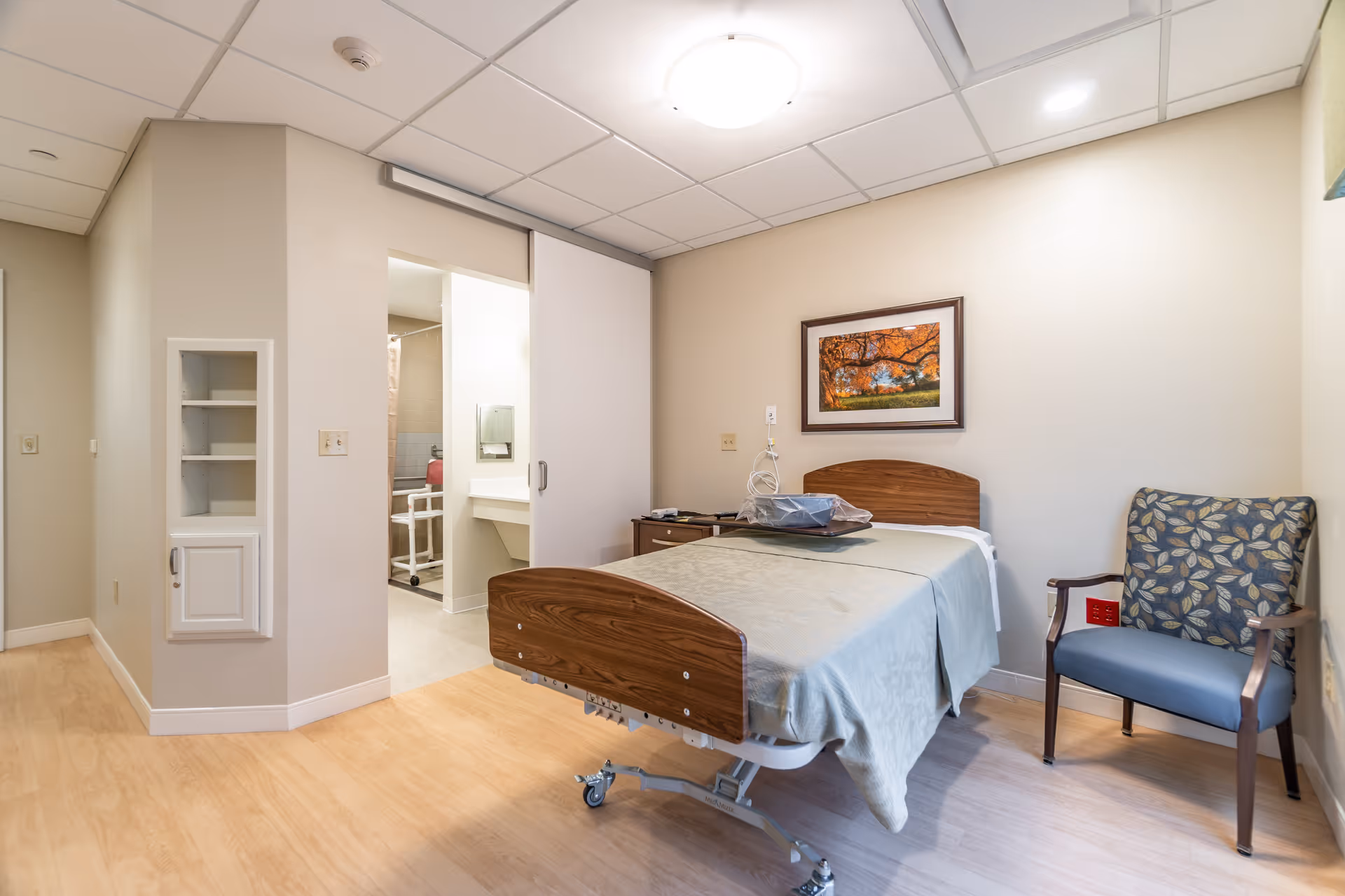 A furnished senior facility bedroom with a hospital-style bed, bedside table, armchair, and an open doorway to a bathroom.