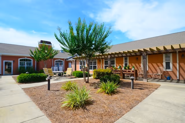 Outdoor courtyard area of a senior living facility with a central garden bed containing small trees and shrubs, surrounded by paved walkways. There are patio chairs and tables on the left side and rocking chairs under a pergola on the right side, with a clear blue sky overhead.
