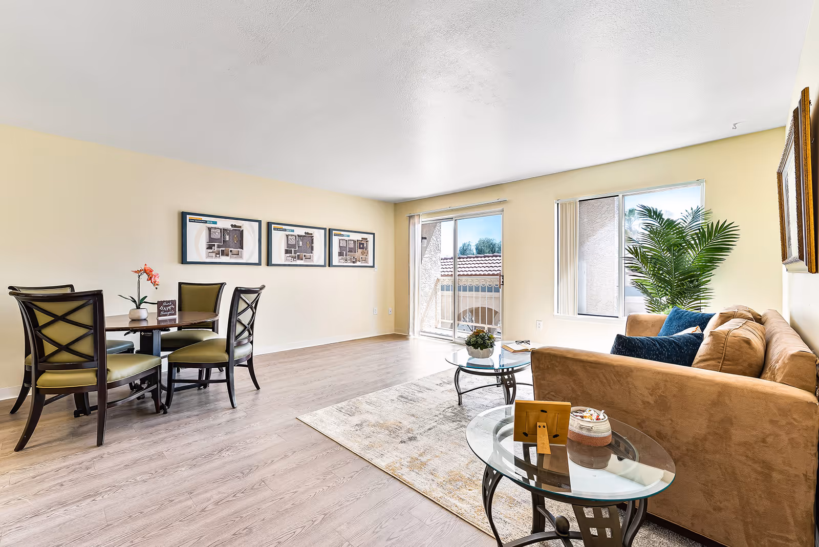 Bright furnished living room and dining area with a sofa, glass coffee tables, a round dining table with four chairs, and a sliding glass door to a balcony.