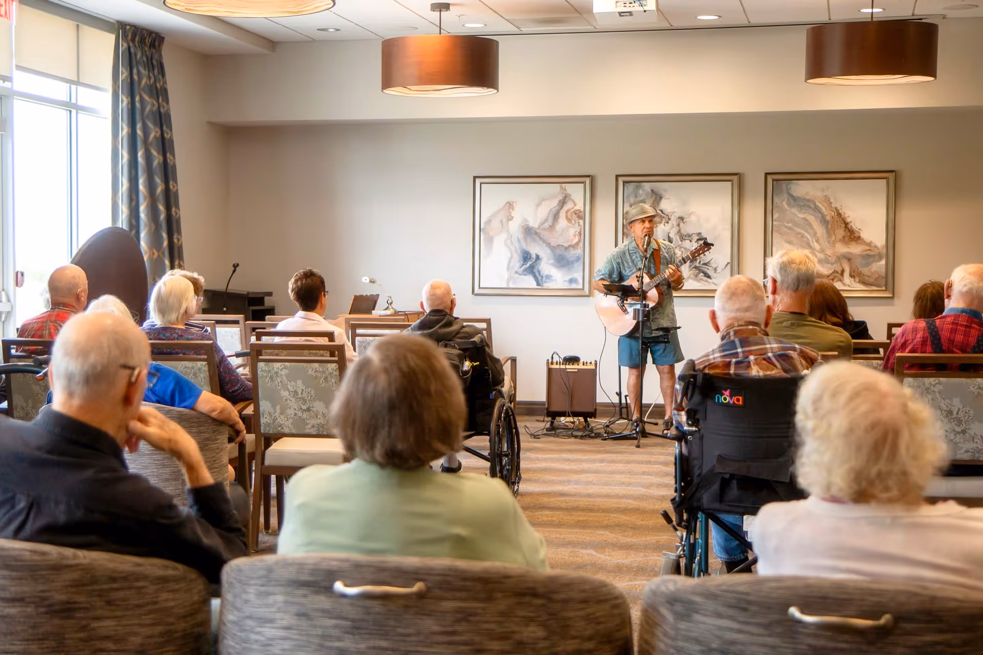 A group of elderly people seated in a room watching a man playing guitar and singing. The room has large windows with curtains, modern ceiling lights, and three abstract paintings on the wall behind the performer.