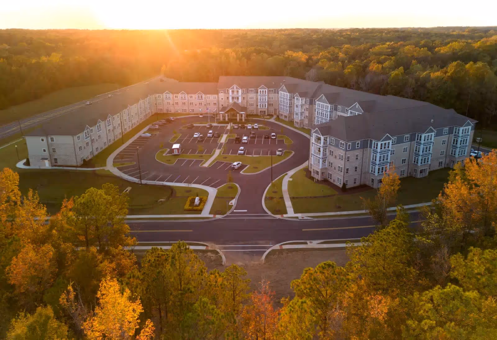Aerial view of a large senior living facility building named The Crossings at North River, surrounded by trees with autumn foliage during sunset. The building is U-shaped with a parking lot in the center and a road in front.
