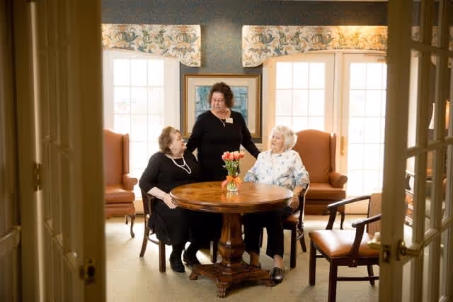 Three women sitting and standing around a round wooden table with a vase of flowers in a well-lit room with large windows and floral valances. Two women are seated in armchairs while the third woman stands between them, engaging in conversation.