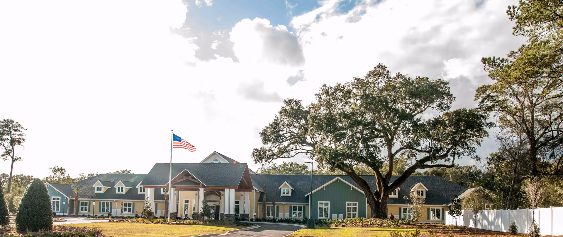 Front exterior view of a senior living facility named YOURLife of Pensacola with a large American flag on a flagpole, a driveway leading to the entrance, and large trees surrounding the building under a partly cloudy sky.