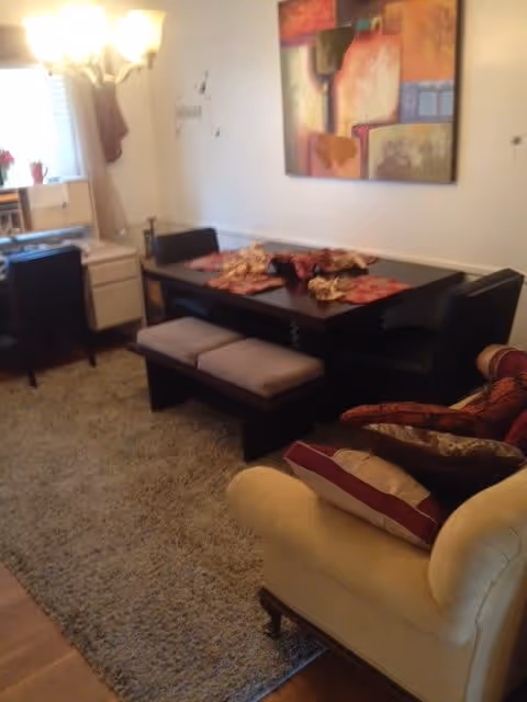 Cozy living area with a dark wood dining table and bench, upholstered armchair, shag rug, floor lamp, and large abstract wall art.