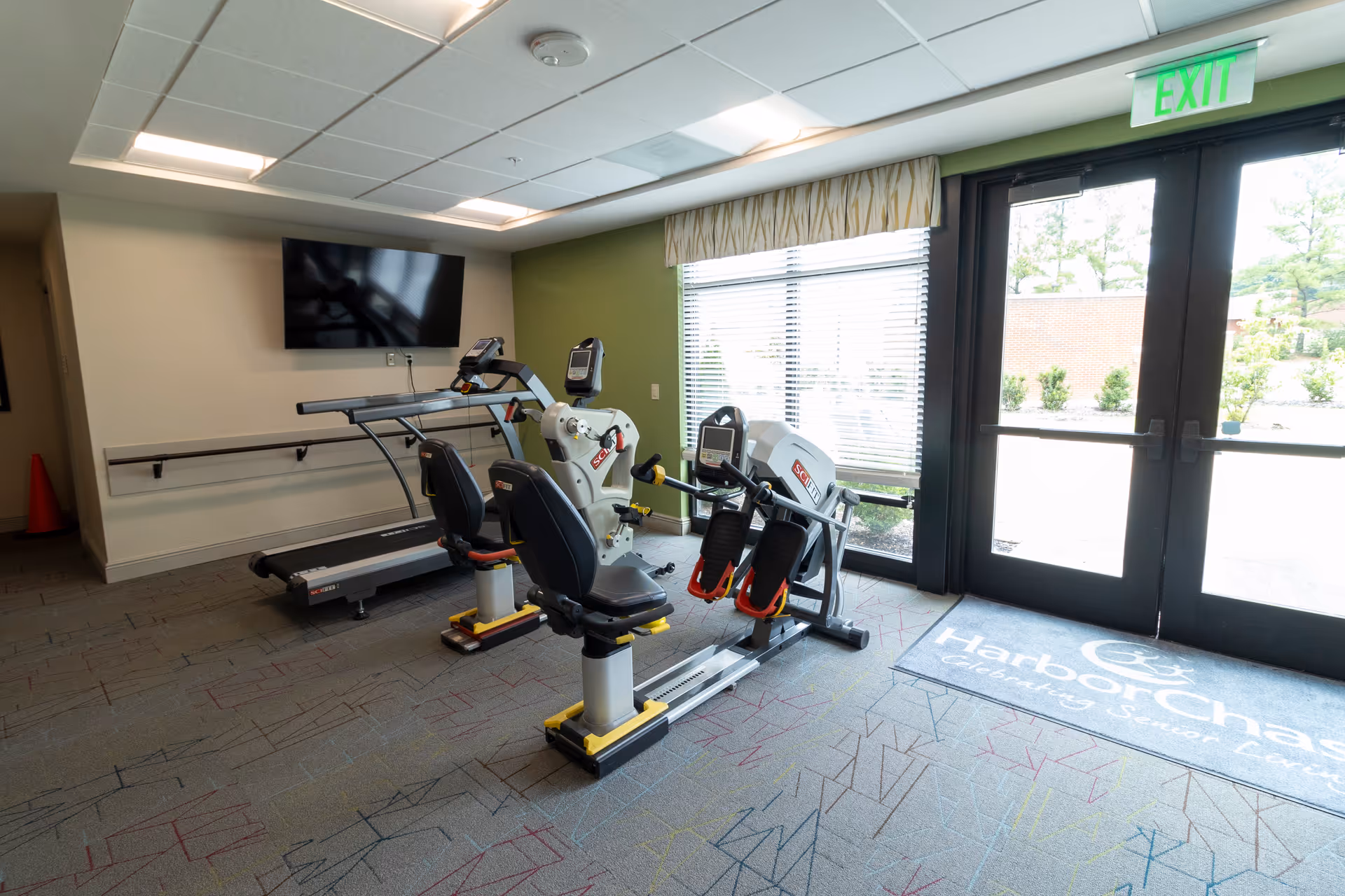 Small exercise room with two recumbent bikes, a treadmill, a wall-mounted TV, and glass exit doors letting in natural light.