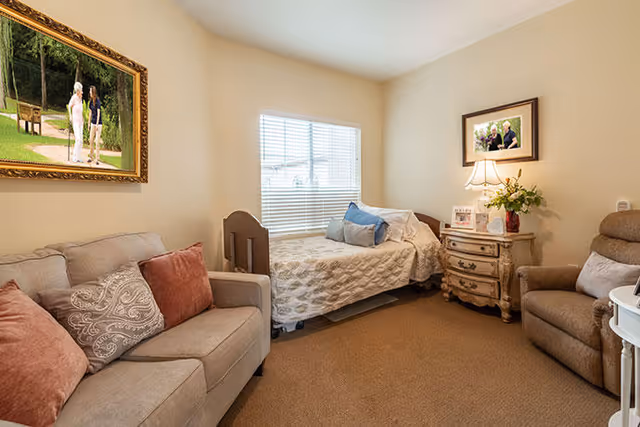 A cozy living space with a beige sofa adorned with patterned and solid pillows, a single bed with a quilt and pillows near a window with blinds, a wooden nightstand with a lamp, framed photos, and a flower vase, and a comfortable recliner chair. The walls are decorated with framed pictures.