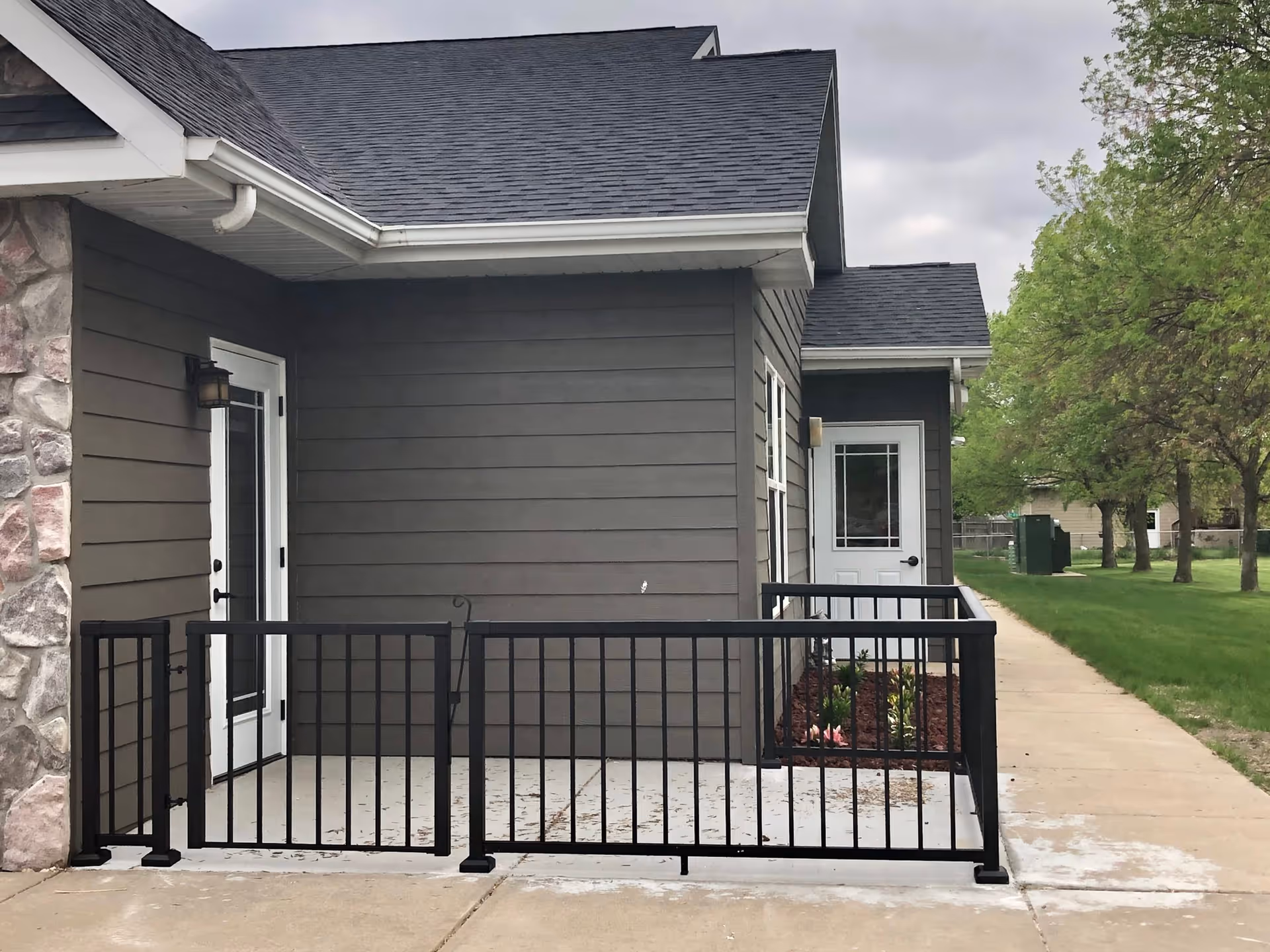 Exterior view of a building corner with gray siding and a stone accent wall. There are two white doors with glass panels, a black metal railing enclosing a small concrete patio area, and a concrete sidewalk leading alongside the building. Green grass and trees are visible in the background under a cloudy sky.