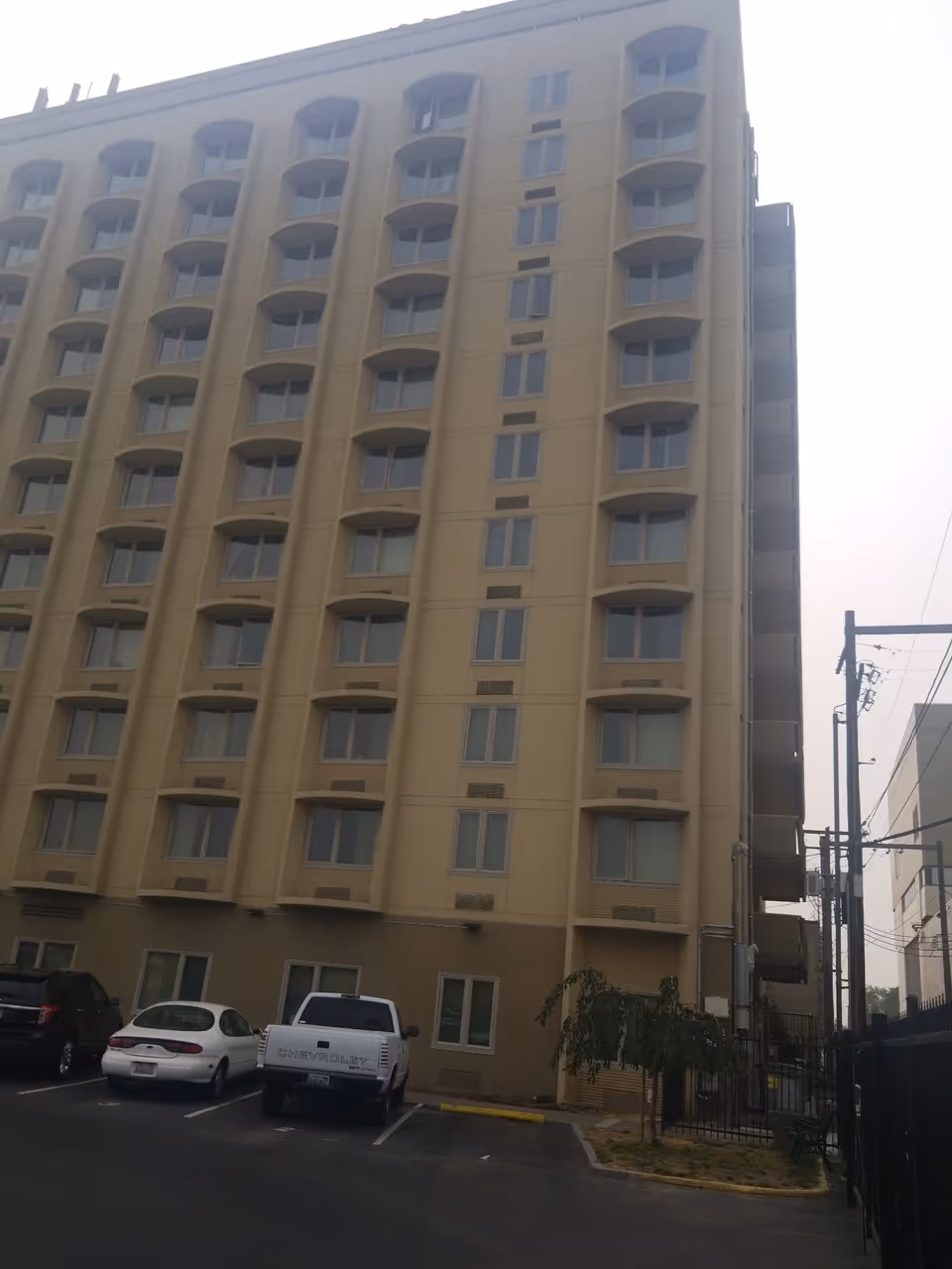Exterior view of a multi-story beige building with multiple windows and a small parking lot in front containing several parked vehicles. There is a small tree and some utility poles visible on the right side.