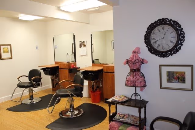 Interior view of a hair salon area with two black salon chairs in front of mirrors and wooden counters. A decorative pink dress form stands on a small table with magazines underneath. A large ornate clock and framed pictures hang on the wall.