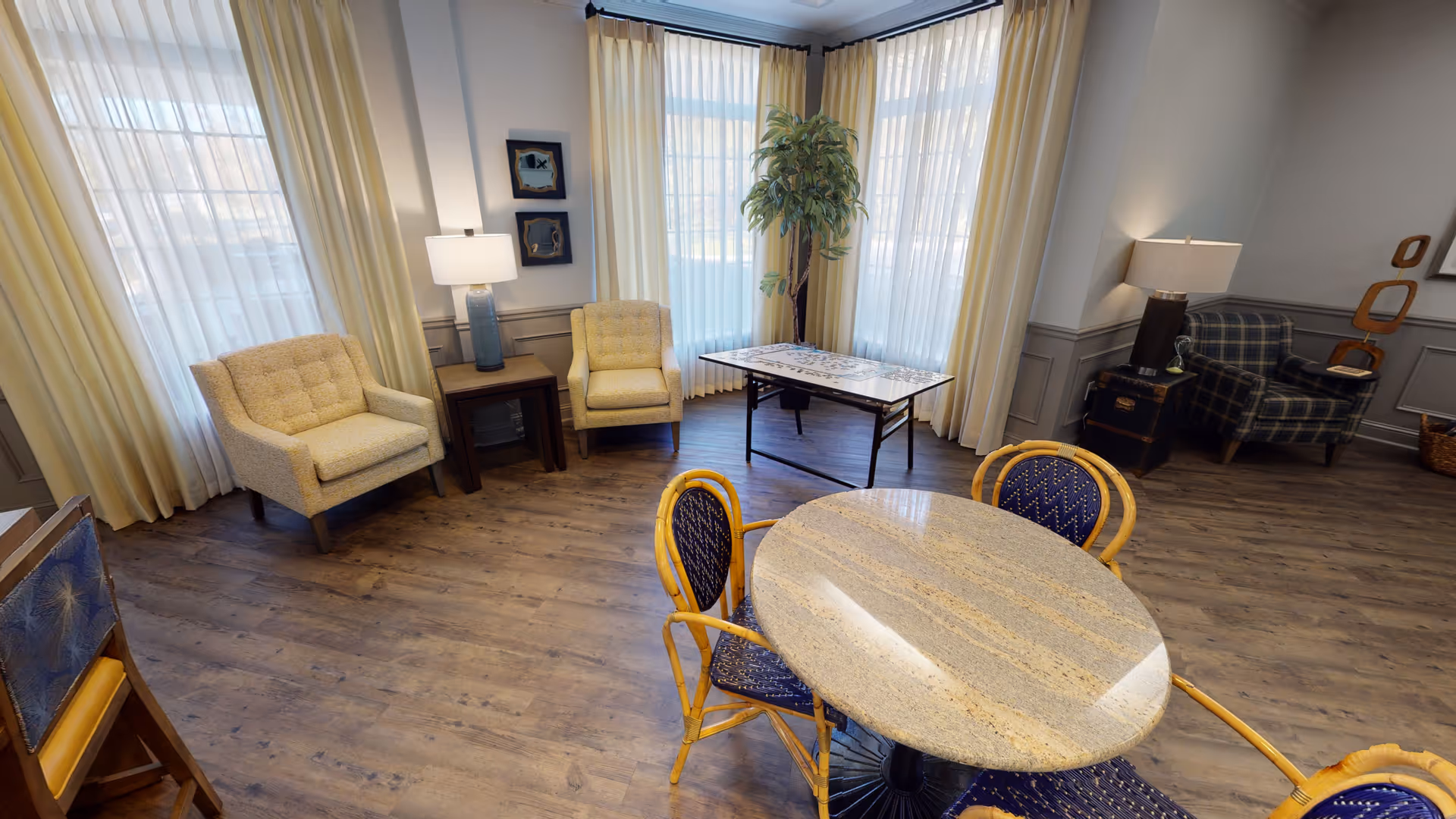 Bright communal lounge with armchairs, a round table and chairs, lamps, and large curtained windows.