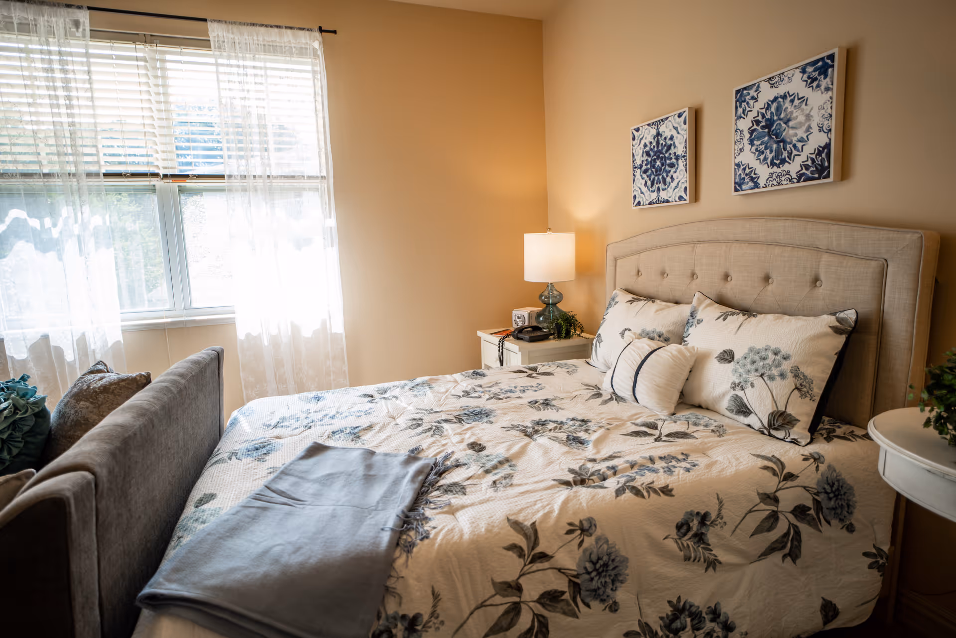 A cozy bedroom with a large bed featuring a floral patterned comforter and multiple pillows. There is a beige upholstered headboard, two framed floral artworks on the wall above the bed, a nightstand with a lamp and a phone, and a window with sheer white curtains letting in natural light.