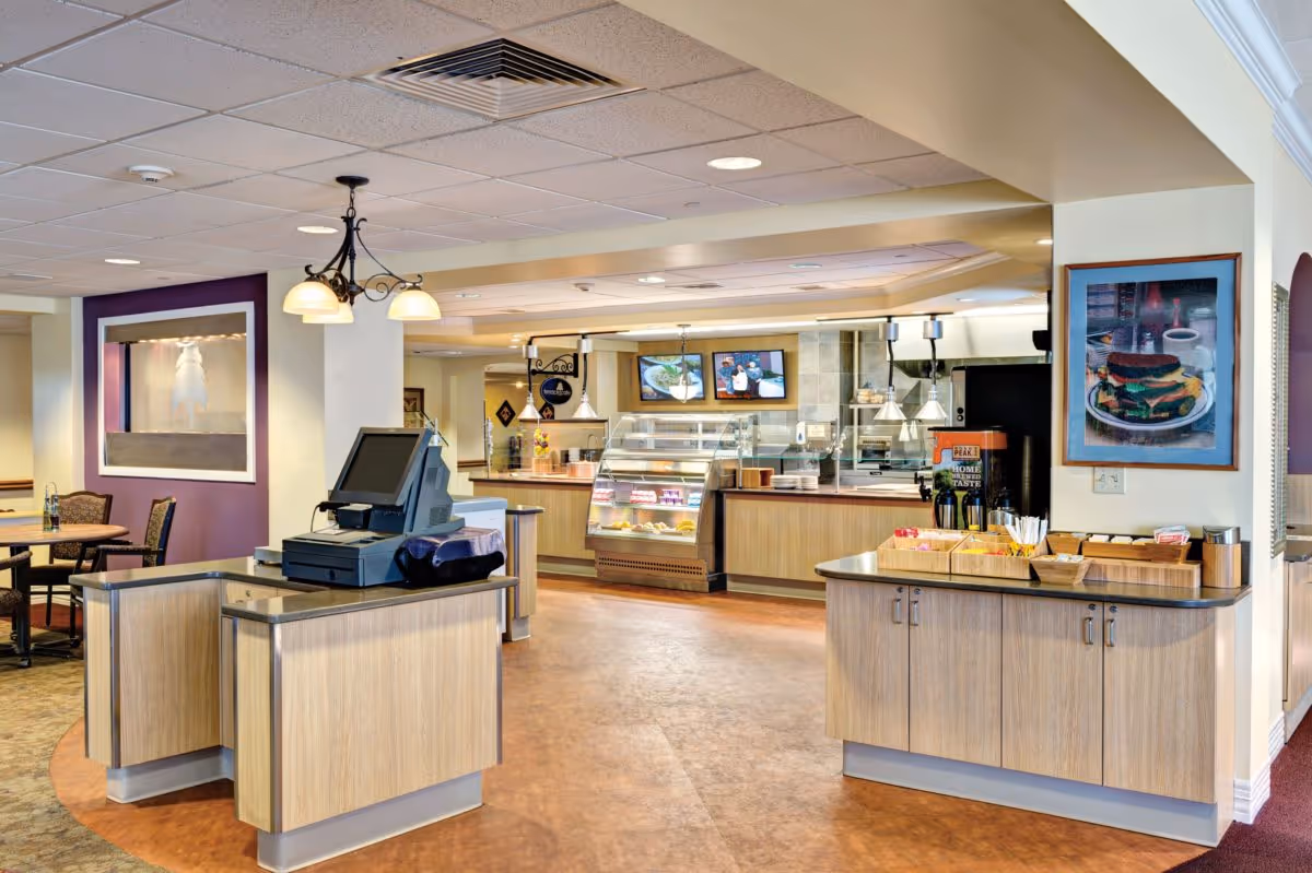 Interior view of a dining/cafe area with serving counters, a register station, seating, and framed artwork.