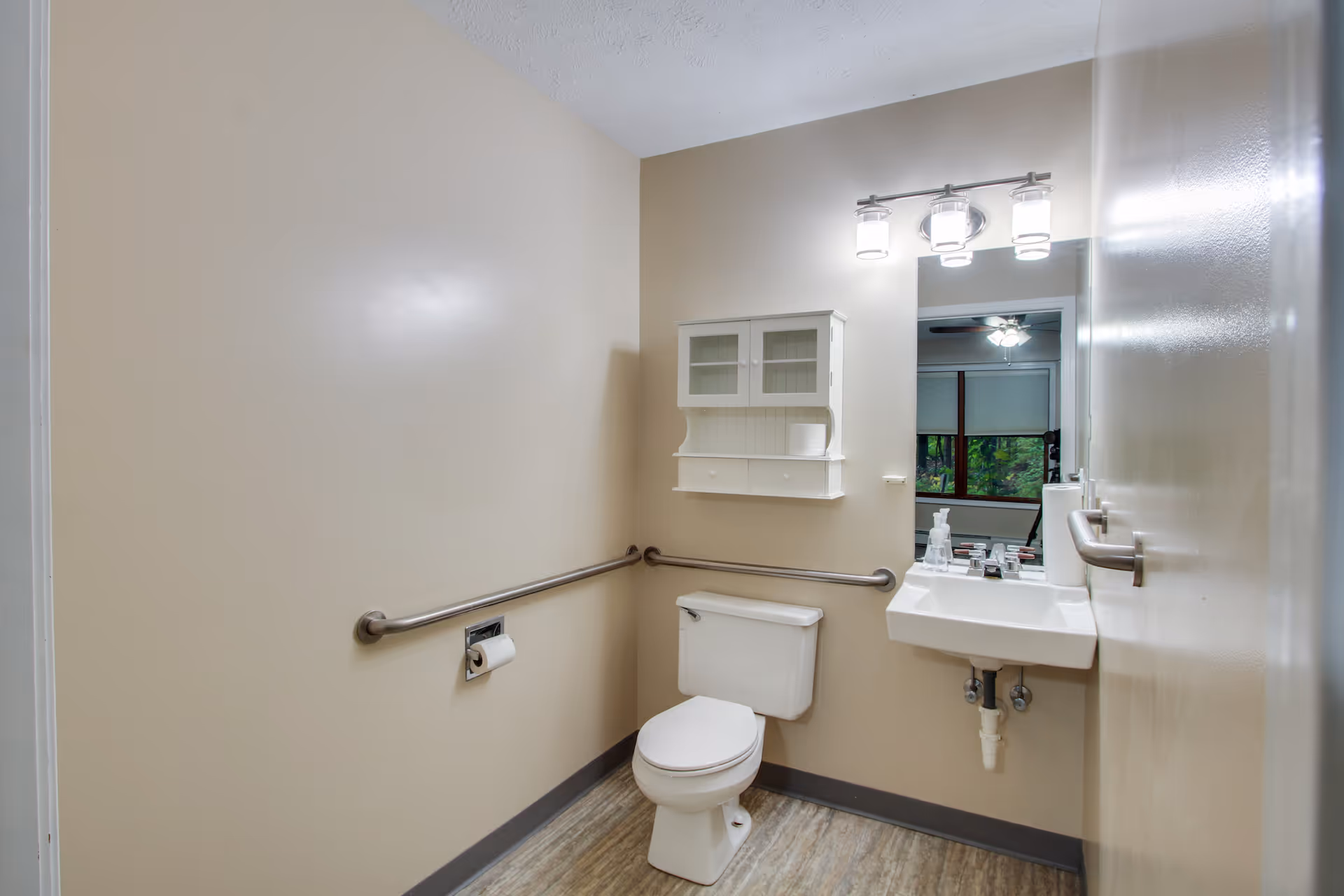 A clean, accessible bathroom with beige walls and wood-look flooring. The room features a white toilet with a grab bar on the wall beside it, a small white wall-mounted cabinet above the toilet, and a white sink with a mirror and three-light fixture above it. There is also a grab bar on the door.