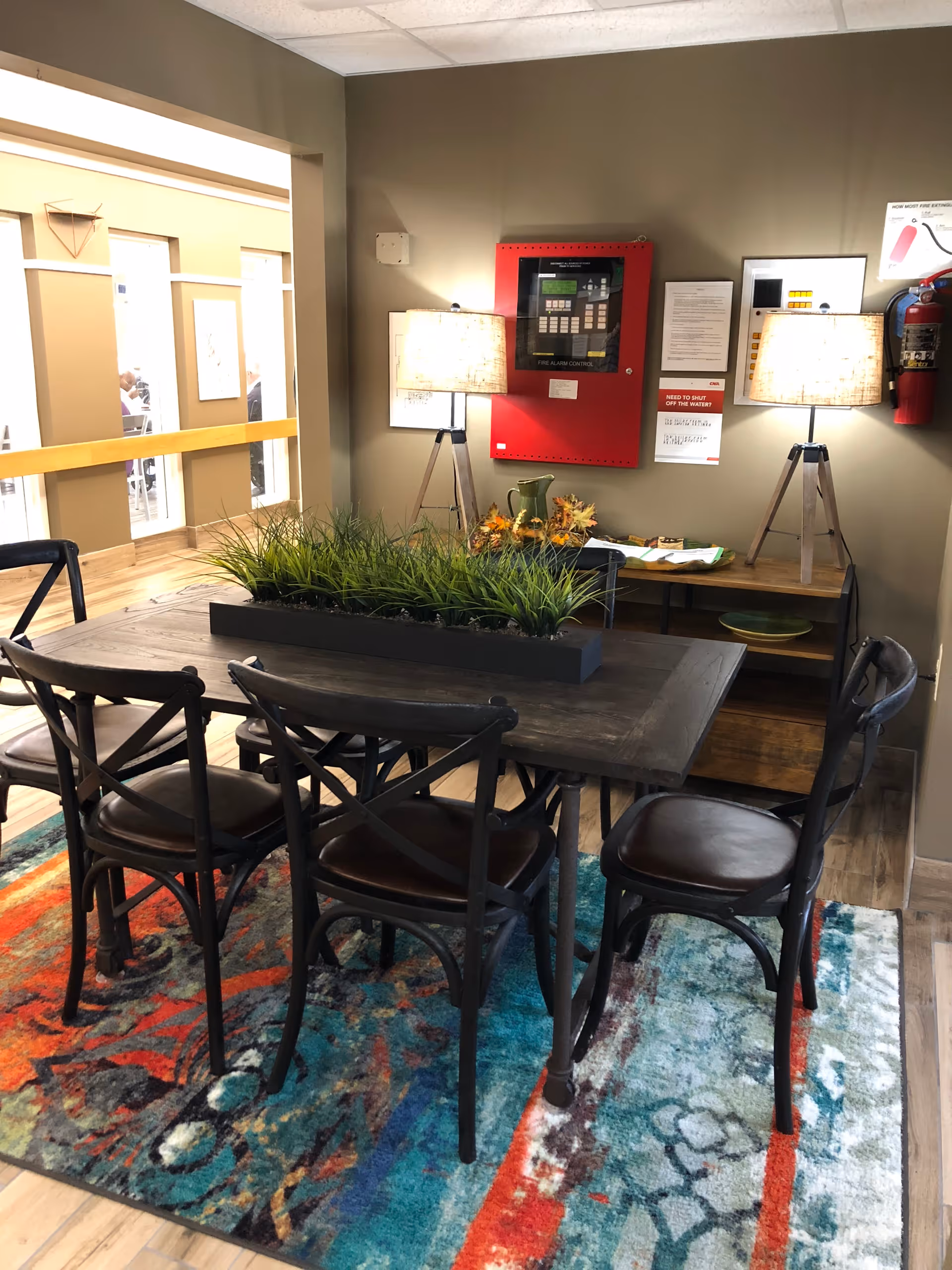 A cozy interior room featuring a wooden table with six dark wooden chairs arranged around it. The table has a rectangular planter with green grass-like plants. The floor is covered with a colorful patterned rug in shades of red, blue, and gray. Against the wall, there is a wooden sideboard with two tripod lamps with beige lampshades, a green pitcher, and some decorative items. A red fire alarm control panel and a fire extinguisher are mounted on the wall above the sideboard.