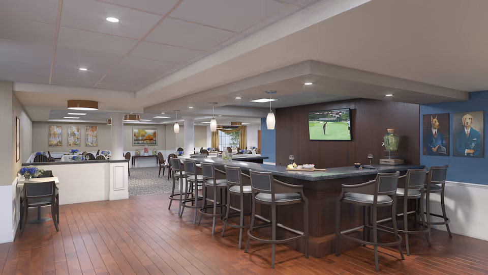 Interior view of a senior living facility dining and lounge area with a large bar counter surrounded by high chairs. A flat-screen TV is mounted on a dark wood wall behind the bar, showing a golf game. The room has wooden flooring, pendant lights hanging above the bar, and several dining tables with chairs in the background. The walls are decorated with paintings, including two portraits of dogs dressed in suits.