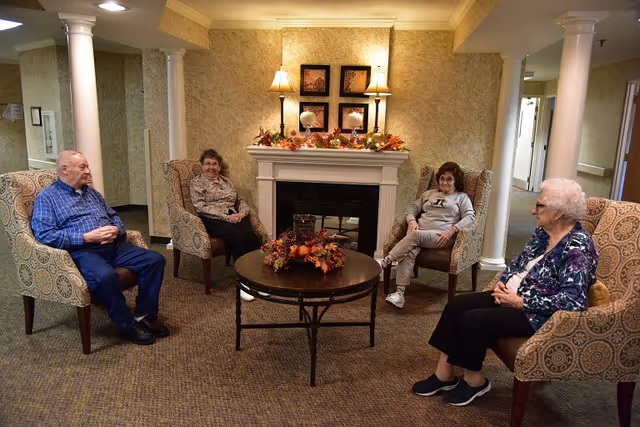 Four elderly people sitting in armchairs arranged in a circle around a round coffee table with a fall-themed centerpiece in a cozy room with a fireplace decorated with autumn leaves and two lamps on the mantel.