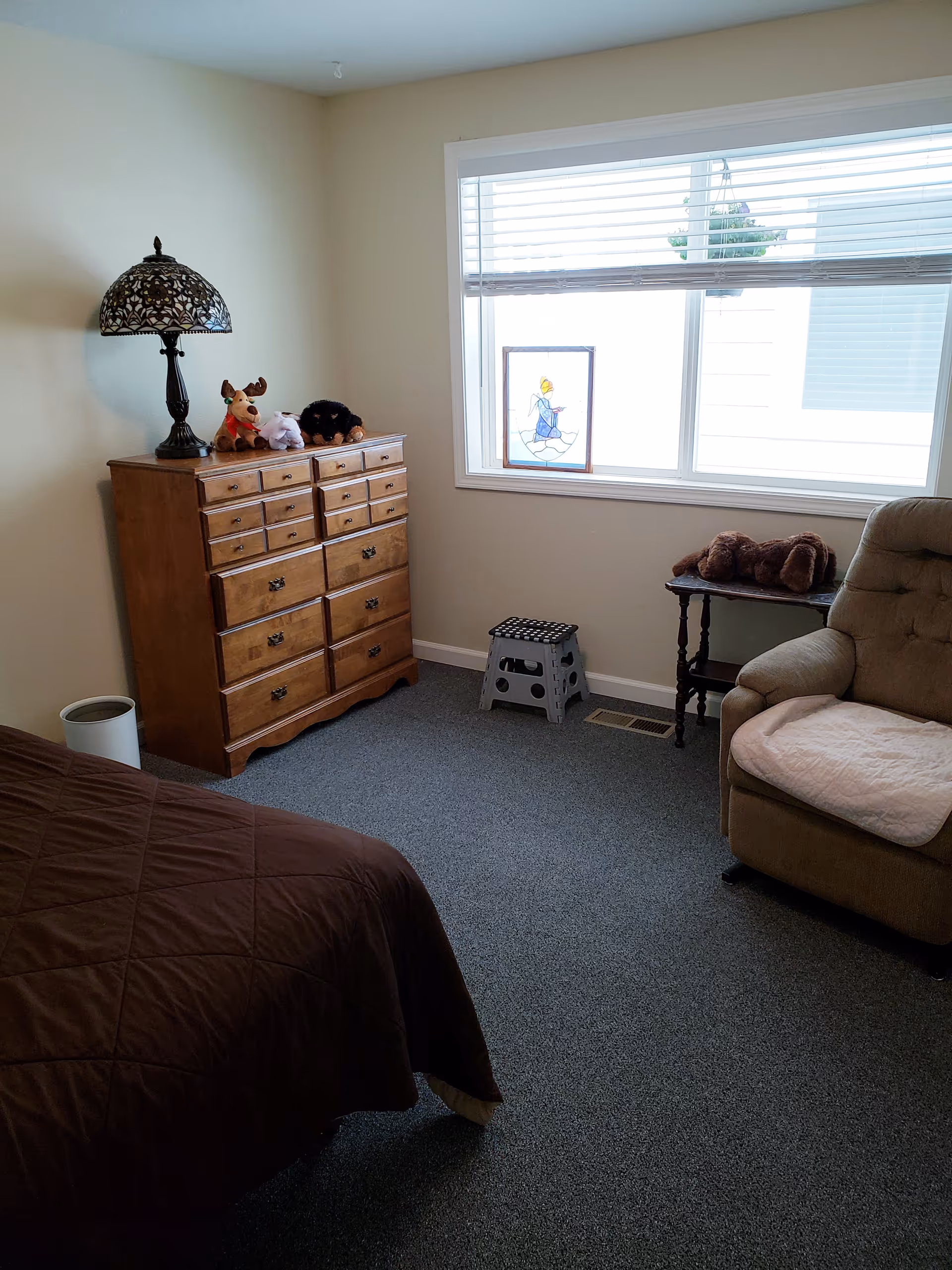 A cozy bedroom with a brown bedspread partially visible in the foreground, a wooden dresser with a decorative lamp and stuffed animals on top, a window with blinds and a stained glass decoration, a small step stool on the floor, a small side table with a stuffed dog, and a cushioned armchair with a light-colored cover.