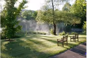 Sunlit grassy lawn with two wooden benches near a misty pond surrounded by trees.