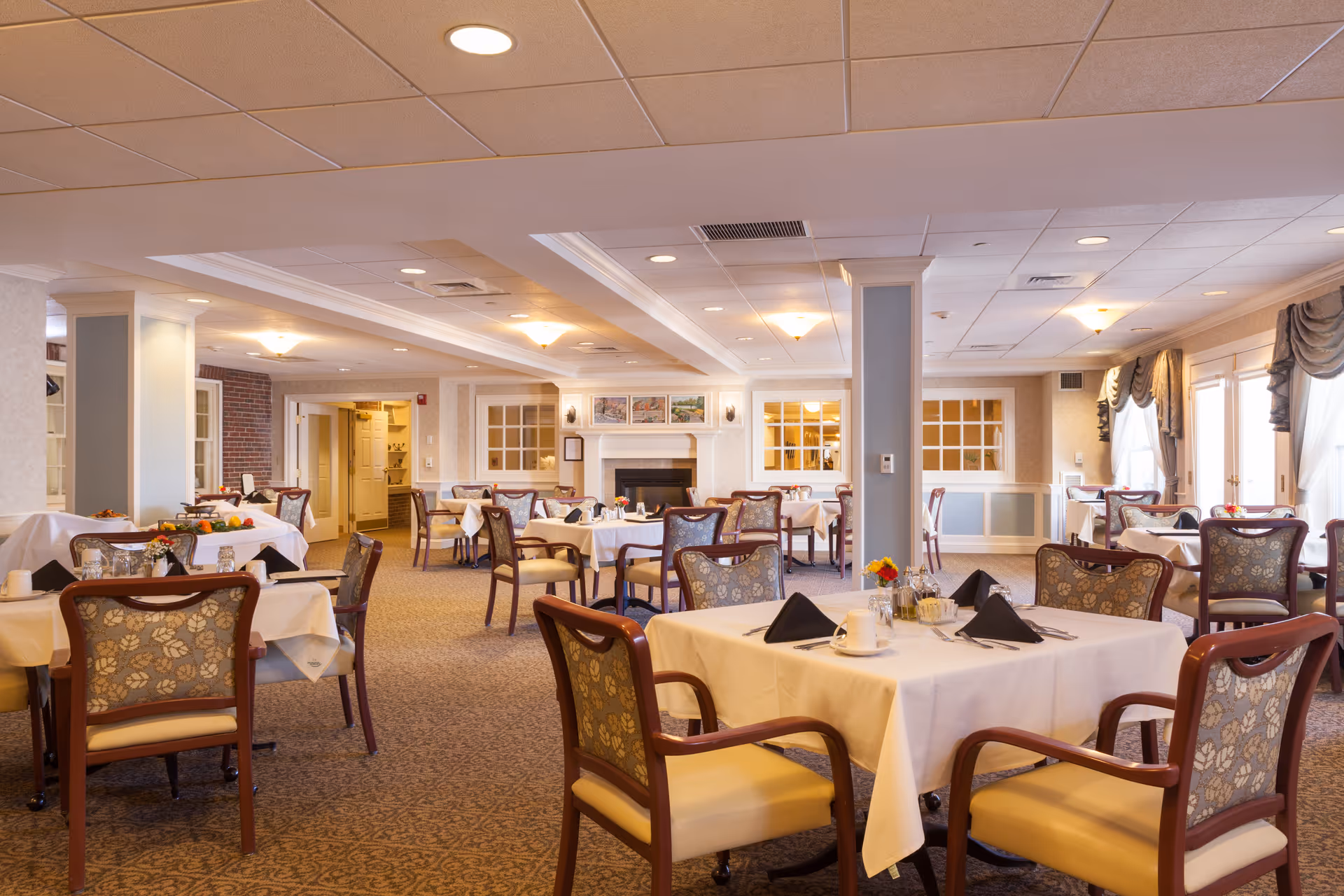 Well-lit dining room with multiple tables set with white tablecloths and chairs in a senior living facility.
