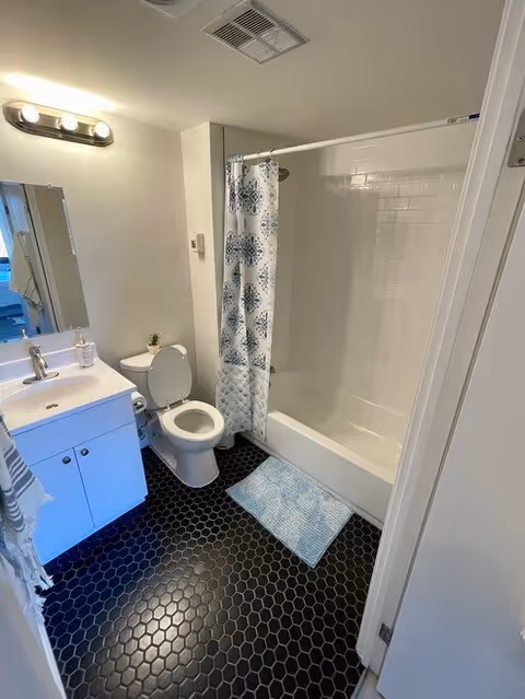 A clean bathroom featuring a white vanity with a sink and faucet, a mirror above the sink, a toilet with the lid open, a bathtub with a white and blue patterned shower curtain, and a light blue bath mat on a black hexagonal tile floor.