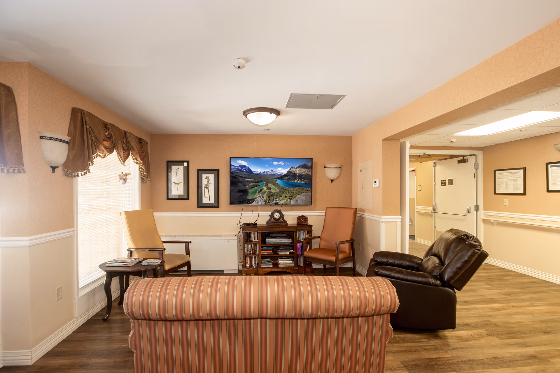 A cozy living room area in Newton Presbyterian Manor with a striped sofa in the foreground, two armchairs, a side table with magazines, a wall-mounted flat screen TV displaying a scenic mountain and lake view, framed artwork on the wall, and warm beige walls with white trim. The room has wood flooring and a ceiling light fixture, with an open doorway leading to a hallway.