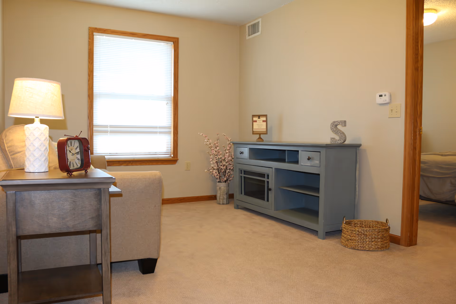 A cozy living room area with beige carpet and walls. There is a beige armchair next to a wooden side table with a white lamp and a red clock on it. A window with blinds is on the left wall. Against the right wall is a blue-gray entertainment console with decorative items on top, including a framed certificate and a letter 'S'. A woven basket sits on the floor near the doorway leading to a bedroom with a visible bed.