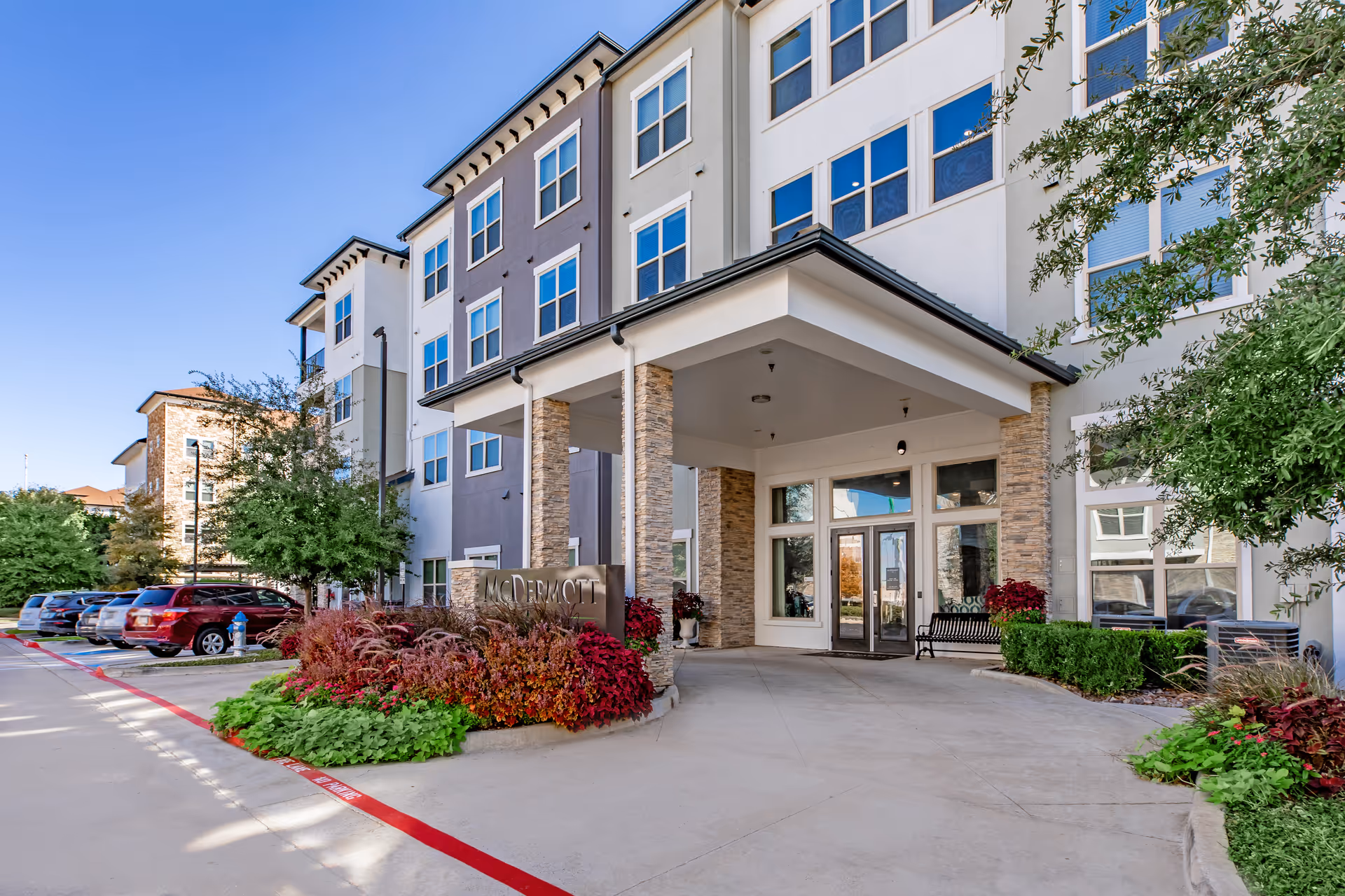 Front entrance of McDermott Crossing senior living building with a covered drive, landscaping, and parked cars.